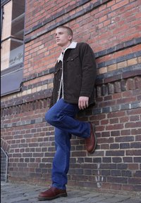 Chaqueta de pana marrón con cuello de borreguillo blanco, combinada con jeans azules y botas de cuero marrón; de pie contra una pared de ladrillo.