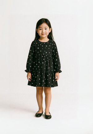 Schwarzes Kleid mit langen Ärmeln, gestuftem Design, silbernem Sternenmuster und elastischen Bündchen. Kombiniert mit schwarzen flachen Schuhen.