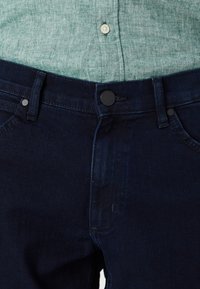 Jean en denim bleu foncé avec boutons, porté avec une chemise texturée vert clair rentrée à l'intérieur.