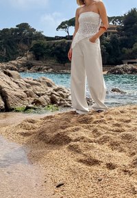 Vit axelbandslös topp med texturerad spetskant, tillsammans med löst sittande krämfärgade byxor, står på sandstrand nära klippig strand och vatten.