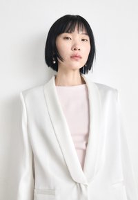 White tailored blazer with a satin finish, notched lapels, and side pockets over a light pink top. Model has short black hair and pearl earrings.