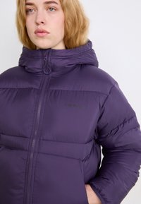 Puhu jakna v temno vijolični barvi, z visokim ovratnikom, zadrgo in stranskimi žepi. Teksturirana, oblazinjena zasnova z logotipom.