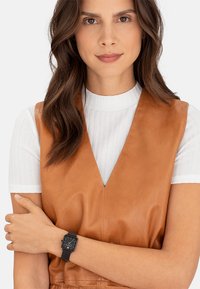 Montre noire carr ée au poignet, avec un cadran minimaliste. Le modèle porte un gilet en cuir tan par-dessus une chemise blanche à côtes.