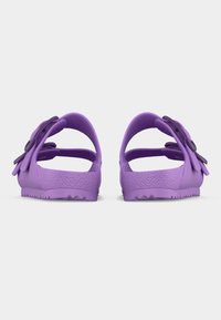 Ζευγάρι μωβ σανδάλια με ανοιχτή φτέρνα, ρυθμιζόμενα λουριά και υφασμένα πέλματα, εμφανιζόμενα από πίσω σε λευκό φόντο.