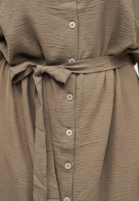 Φόρεμα από ελαφρώς καφέ υφασμένο ύφασμα με ζώνη στη μέση και μία κάθετη σειρά από έξι στρογγυλά μπεζ κουμπιά στο μπροστινό μέρος.