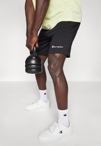 Black shorts with a Champion logo, paired with white sneakers and socks featuring the Champion emblem. Weights held in one hand.