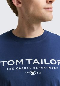 Navy blauwe katoenen t-shirt met witte tekst "TOM TAILOR THE CASUAL DEPARTMENT 1962," met een ronde hals en korte mouwen.