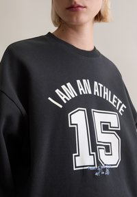 Czarna bluza z białym napisem "JESTEM ATLETĄ" oraz numerem "15". Posiada okrągły dekolt i teksturowany materiał.
