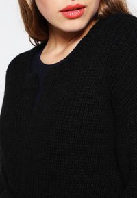 Žena s světlou pletí a hnědými vlasy, oblečená v texturovaném černém svetru a korálovými rtěnkami, zobrazená od krku po ramena.