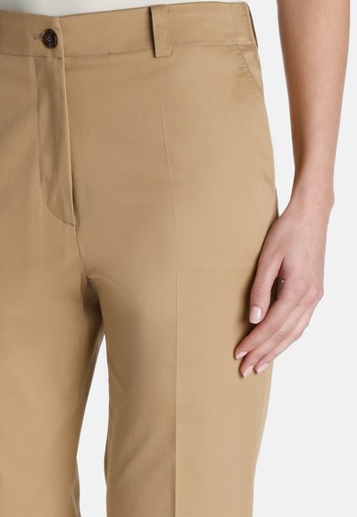 Beige housut, joissa on sileä, teksturoitu pinta, vyötäröllä nappikiinnitys, sivutaskut ja mittatilaustyönä tehty istuvuus.