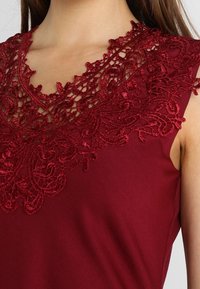 Vínový top s detailním červeným květinovým krajkovým zdobením podél výstřihu do V a hladkou texturou látky. Bez rukávů.