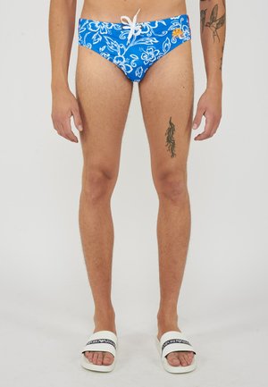 Uomo che indossa slip da bagno blu con motivo floreale e ciabatte bianche Emporio Armani, mostrando i tatuaggi su braccia e parte superiore delle gambe su uno sfondo semplice.