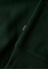 Tela de sudadera verde oscuro con puños acanalados, con una pequeña etiqueta negra que dice "SUPERDRY" en el lateral. Textura suave y costuras limpias.