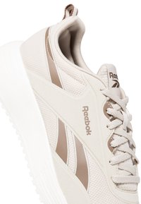 Zapatilla deportiva beige con parte superior de malla texturizada, acentos suaves, rayas marrón claro y suela de goma blanca. Logo de Reebok visible.