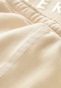 Pantalons joggers beiges en matière texturée, avec une taille élastique et un détail de logo sur le côté en lettres en relief.