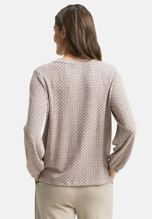 Donna con lunghi capelli castani che indossa un top beige a maniche lunghe con un motivo e pantaloni chiari, con le mani nelle tasche, di spalle.