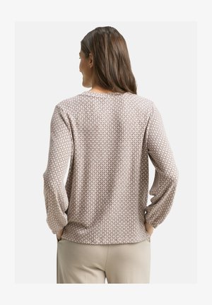 Femme aux longs cheveux bruns portant un haut à manches longues beige à motifs et un pantalon clair, les mains dans les poches, de dos.