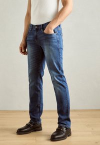 Jeans blu con una vestibilità slim, realizzati in denim. Presentano tasche anteriori, una chiusura con bottone e una finitura leggermente sbiadita. Indossati con mocassini neri.