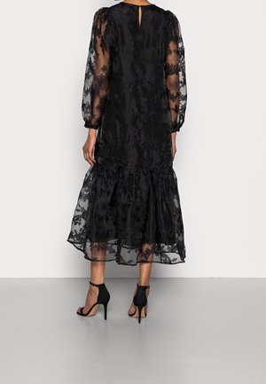 Czarna midi sukienka z przezroczystymi rękawami w kwiatowy wzór oraz warstwową spódnicą, w zestawieniu z czarnymi sandałami na wysokim obcasie z paskiem wokół kostki, widziana od tyłu.