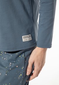 Blauwe shirt met lange mouwen gemaakt van zachte stof, met een rechthoekig label met tekst. Gecombineerd met patroonbroeken met kleine gekleurde vormen.