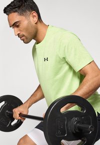 Αθλητική μπλούζα ανοιχτού πράσινου χρώματος με υφή, με μαύρο λογότυπο, που φοριέται από άτομο που σηκώνει μπάρα με βάρη. Απλός στρογγυλός λαιμός.