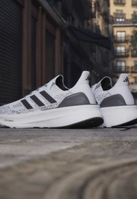 Paire de chaussures de course Adidas Ultraboost blanches et noires placées dans une rue de la ville avec des bâtiments urbains en arrière-plan.