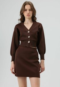 Bruin gebreid tweedelig outfit bestaande uit een cropped vestje met V-hals en drie grote knopen, gecombineerd met een bijpassende aansluitende rok.