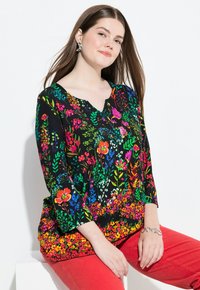 Schwarzes Oberteil mit Blumenmuster und leuchtend bunten Blumen, V-Ausschnitt und Dreiviertelärmeln, kombiniert mit roten Hosen.