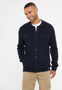 Námořnický modrý pletený cardigan s knoflíkovým zapínáním, dvěma náprsními kapsami a žebrovanými manžetami. Nositý přes bílé tričko a khaki kalhoty.