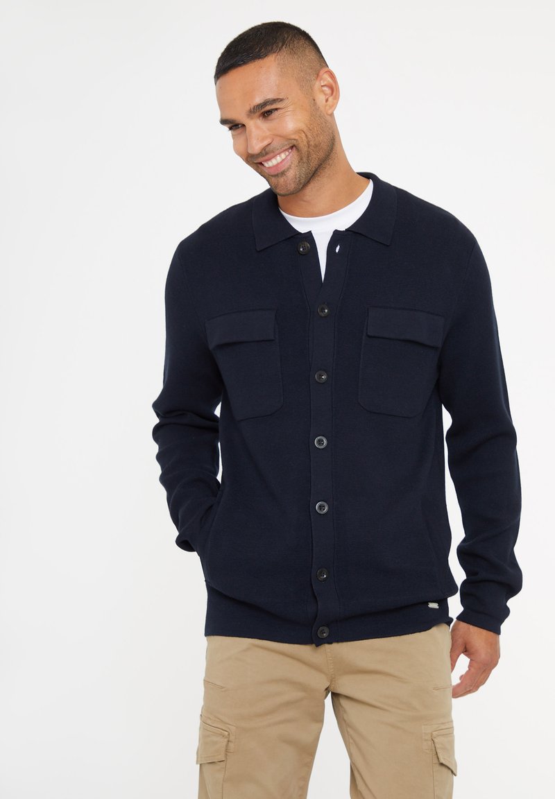 Námořnický modrý pletený cardigan s knoflíkovým zapínáním, dvěma náprsními kapsami a žebrovanými manžetami. Nositý přes bílé tričko a khaki kalhoty.