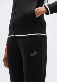 Μαύρο μπουφάν με φερμουάρ και λευκές λεπτομέρειες, μαζί με μαύρη φόρμα με λογότυπο Puma. Το υλικό φαίνεται μαλακό, με λεία υφή.