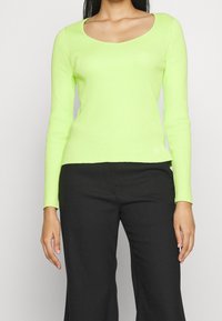 Kvinna som bär en limegrön ribbad långärmad topp med rundad halsringning och svarta byxor mot en enfärgad ljus bakgrund.