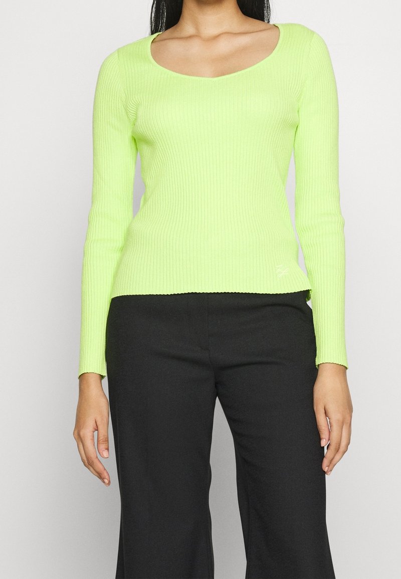 Kvinna som bär en limegrön ribbad långärmad topp med rundad halsringning och svarta byxor mot en enfärgad ljus bakgrund.