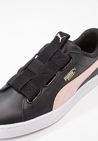 Baskets en cuir noir avec une bande latérale en suède rose, une sangle élastique et une semelle en caoutchouc blanc texturé. Accents dorés du logo sur le côté.
