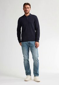 Marineblaues Langarm-Poloshirt mit Kragen, kombiniert mit hellblauen Jeans und weißen Turnschuhen. Glatter Stoff, figurbetontes Design und minimalistische Details.