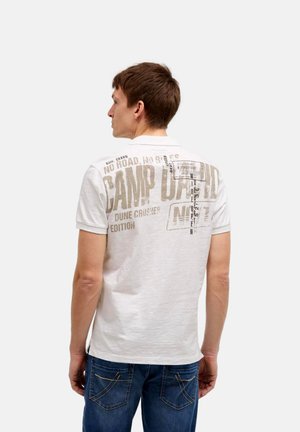 Man wearing white short-sleeve polo shirt with large faded beige text and graphics on back, paired with blue jeans, facing away.