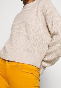 Maglione beige lavorato a maglia con polsini e orlo a coste, abbinato a pantaloni giallo brillante con tasche frontali e dettagli metallici.