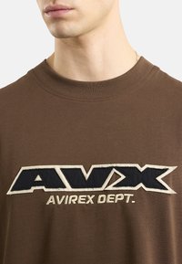 Brązowy bawełniany t-shirt z dużym kremowym i granatowym logo z napisem "AVX AVIREX DEPT." Z ribbowanym dekoltem w kształcie serek i miękką fakturą.