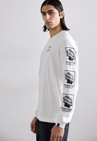 White long-sleeve t-shirt with printed black graphics on both sleeves, featuring a logo design and text. Regular fit with ribbed cuffs.