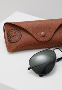 Ochelari de soare aviator negri cu lentile întunecate, expuși alături de o husă din piele maro texturată, cu un logo circular și închidere tip clapetă.