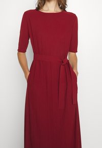 Ein knielanges Kleid in Crimson mit kurzen Ärmeln, strukturiertem Stoff, V-Ausschnitt und einem gebundenen Taillengürtel. Es hat zwei seitliche Taschen.