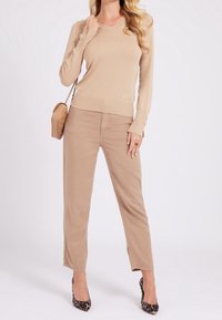 Beżowy sweter z długim rękawem i dekoltem w serek, z dzianinową fakturą i logo marki. W połączeniu z jasnobeżowymi spodniami i czarnymi szpilkami w panterkę.