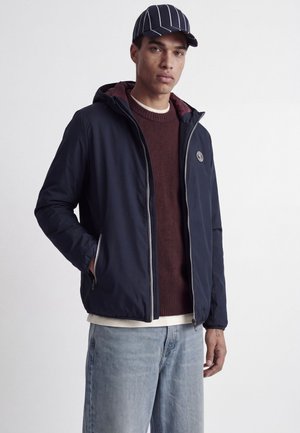 Jeune homme portant une veste zippée bleu marine, un pull bordeaux, un jean bleu clair et une casquette bleu marine à rayures blanches, debout sur un fond blanc.