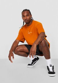 Tricou din bumbac portocalie cu un logo alb, asortat cu pantaloni scurți negri și pantofi de baschet negri. Modelul este în poziția ghemuit, cu brațele sprijinite pe genunchi.