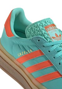 Turkizni čevlji z oranžnimi poudarki. Ponašajo se s tremi oranžnimi črtami, teksturiranim jezikom in zlatim logotipom "Gazelle". Guma podplat.