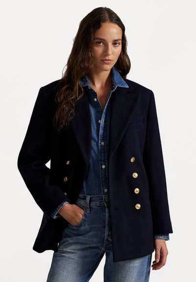 Blazer en mélange de laine navy avec des boutons dorés, présentant un col à revers structuré, porté sur une chemise en denim et un jean, affichant une coupe ajustée.