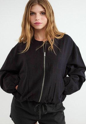 Młoda kobieta z długimi blond włosami, ubrana w czarną kurtkę na suwak z elastycznymi mankietami, z rękami w kieszeniach na białym tle.