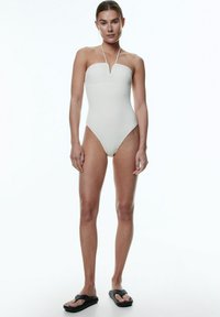 Maillot de bain une pièce blanc texturé sans bretelles avec un décolleté en V ; le modèle porte des tongs noires ; design minimaliste, lignes épurées.