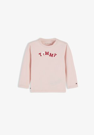 Jasnoróżowa bluzka z długim rękawem dla malucha z napisem "TOMMY" czerwonymi literami na piersi oraz małym logo Tommy Hilfiger na mankiecie rękawa.