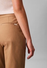 Tuva personāža, kas ģērbusies bēša krāsas biksēs un baltā bezapkaklīšu topā, ar labo roku atslābinātu pie sāniem pelēkā fonā.